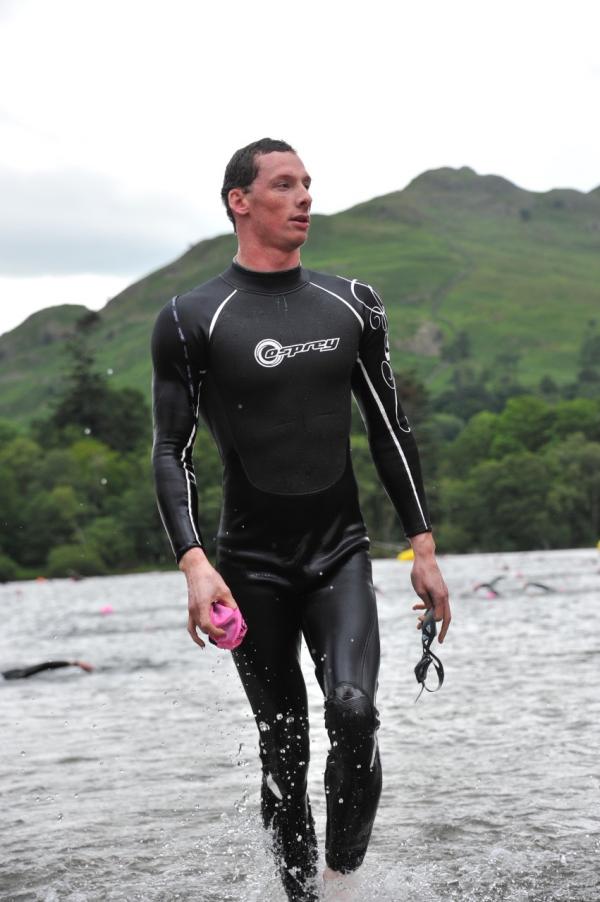 A man emerges from a lake in a swimming suit, holding goggles