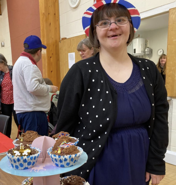 A lady stands in a Union Jack hat next to cakes she has made